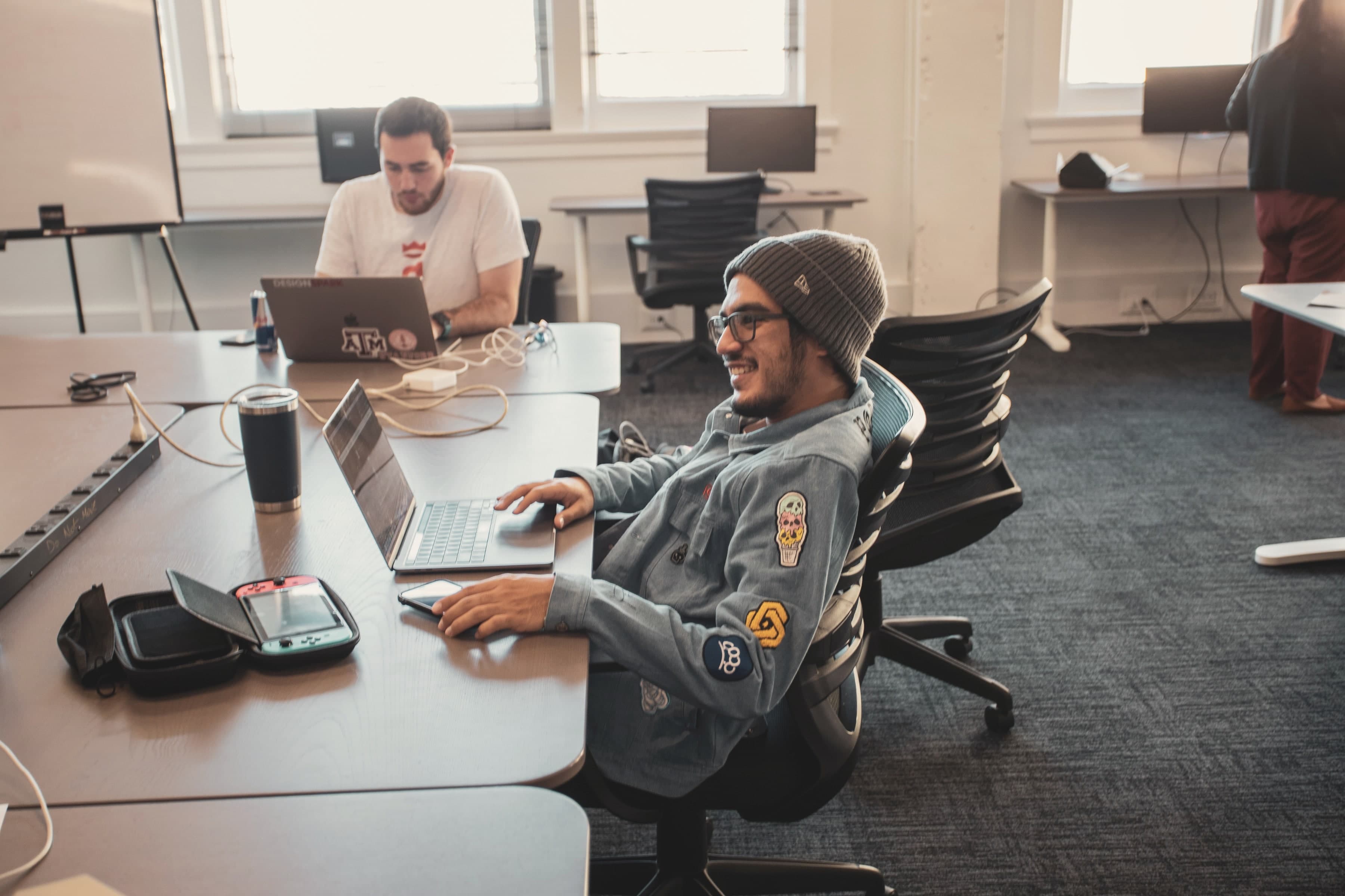 Two members working at laptops on the Geekdom floor, one smiling at his screen, 2021.