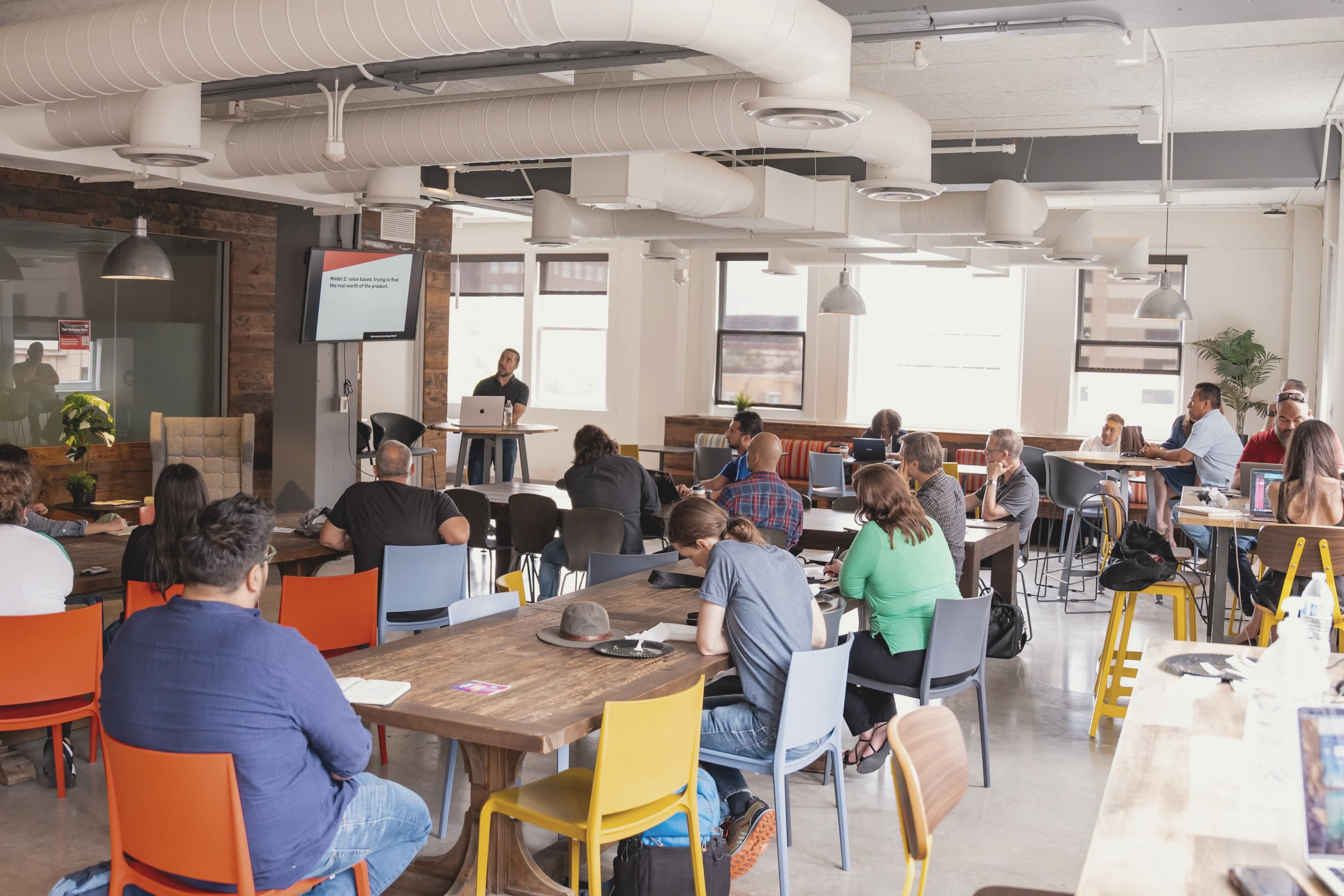 The third floor mid-session: a full room at long wooden tables and orange and yellow chairs, a speaker at the front, windows along the far wall.